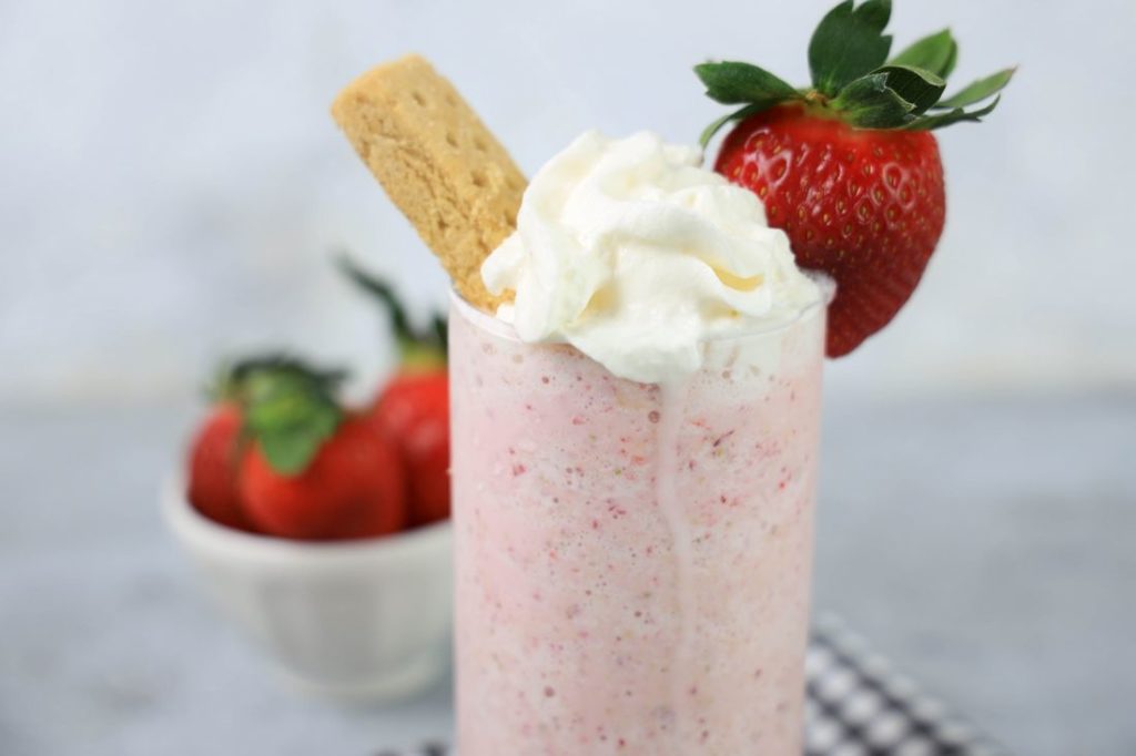 Strawberry Shortcake Shake in a glass with strawberry on top on a gray plaid napkin on a concrete backdrop