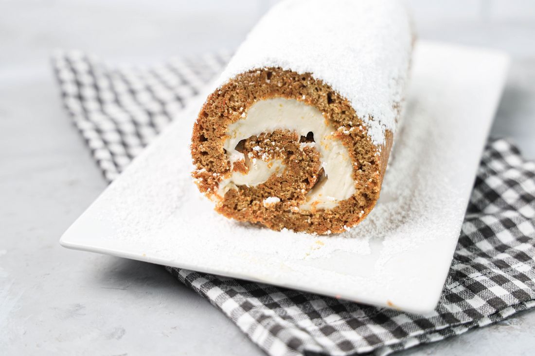 Carrot Cake Roll on a white plate dusted with powdered sugar with a gray plaid napkin and concrete backdrop