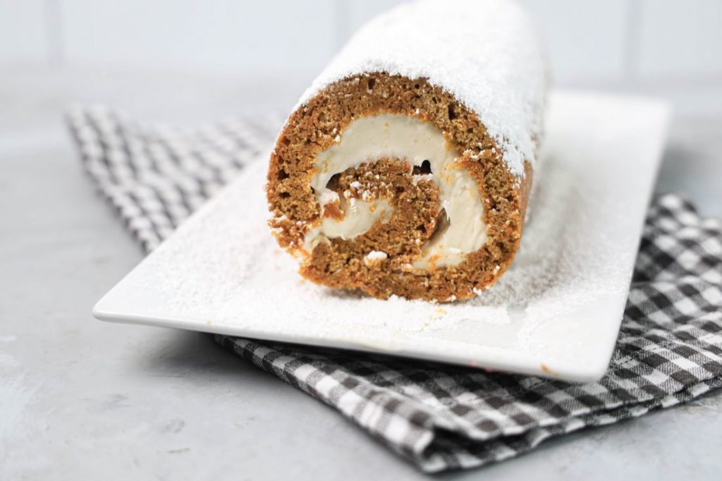 Carrot Cake Roll on a white plate dusted with powdered sugar with a gray plaid napkin and concrete backdrop