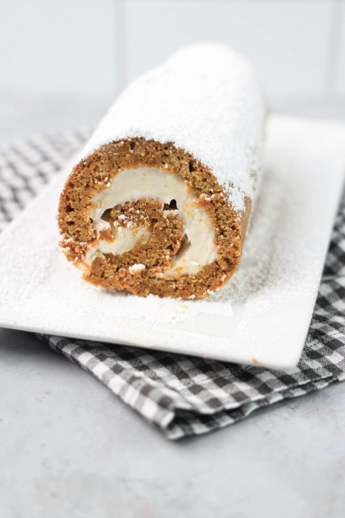 Carrot Cake Roll on a white plate dusted with powdered sugar with a gray plaid napkin and concrete backdrop