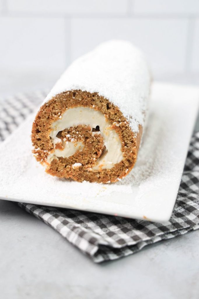 Carrot Cake Roll on a white plate dusted with powdered sugar with a gray plaid napkin and concrete backdrop