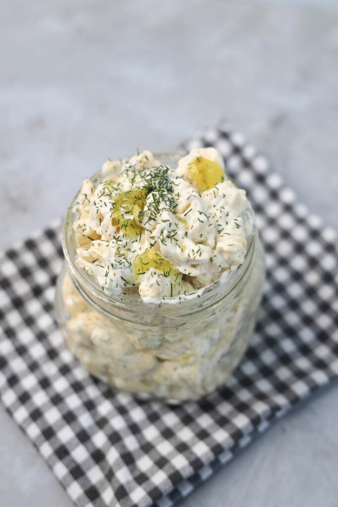 Dill pickle pasta salad in a clear mason jar on a grey plaid napkin on a concrete backdrop