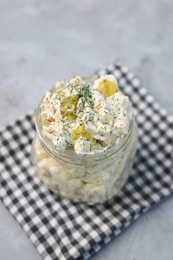 Dill pickle pasta salad in a clear mason jar on a grey plaid napkin on a concrete backdrop