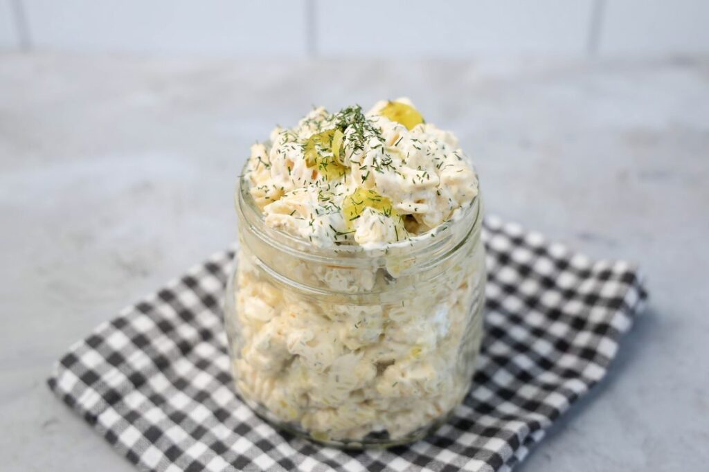 Dill pickle pasta salad in a clear mason jar on a grey plaid napkin on a concrete backdrop
