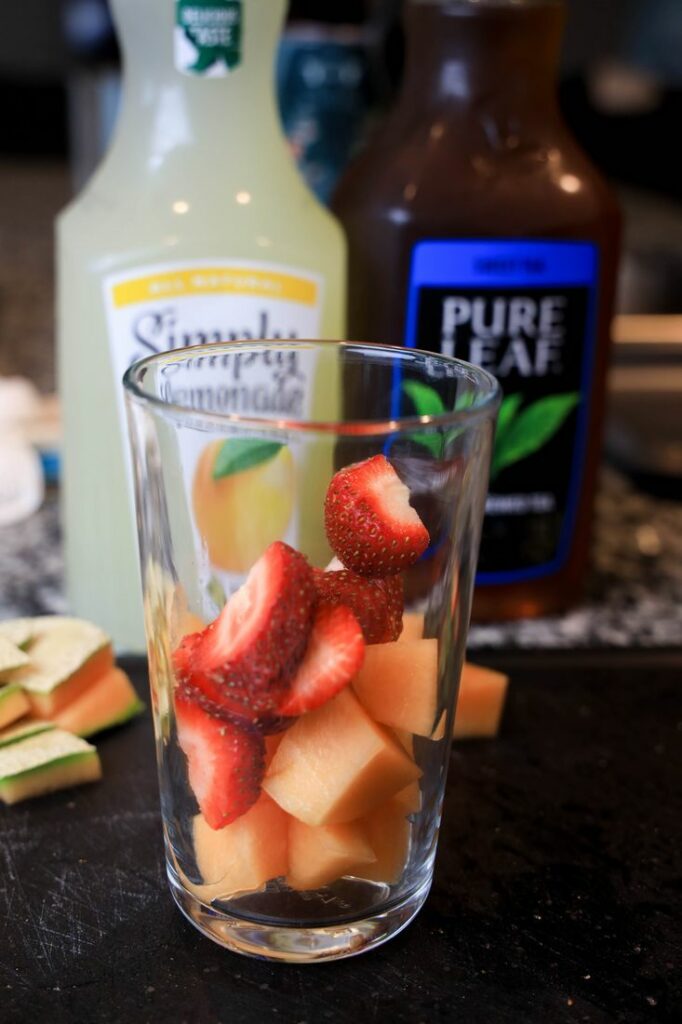 Strawberries and cantaloupe in a glass with lemonade and tea behind it.