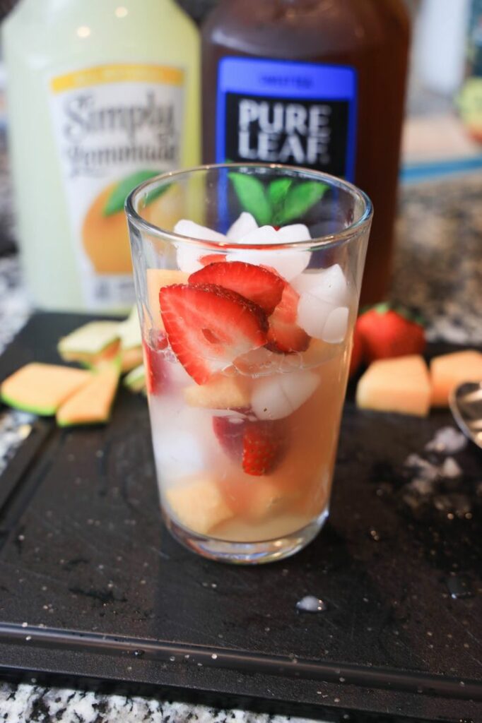 Strawberries and cantaloupe in a glass with lemonade and tea behind it.