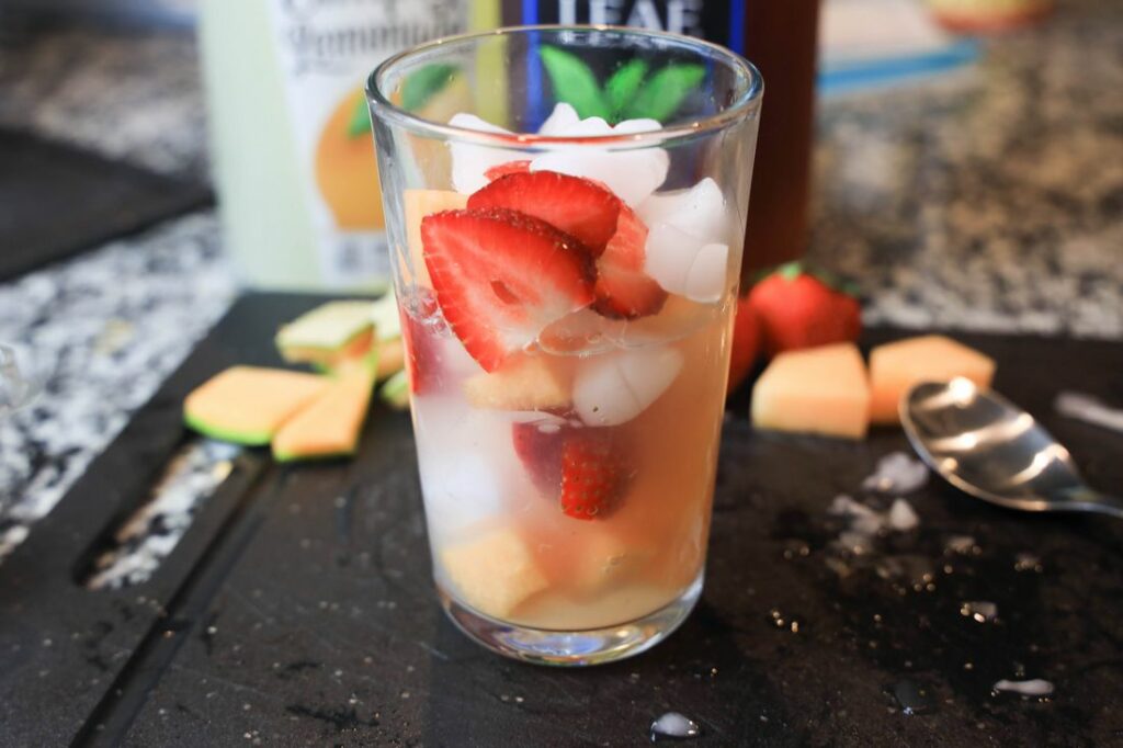 Strawberries and cantaloupe in a glass with lemonade and tea behind it.