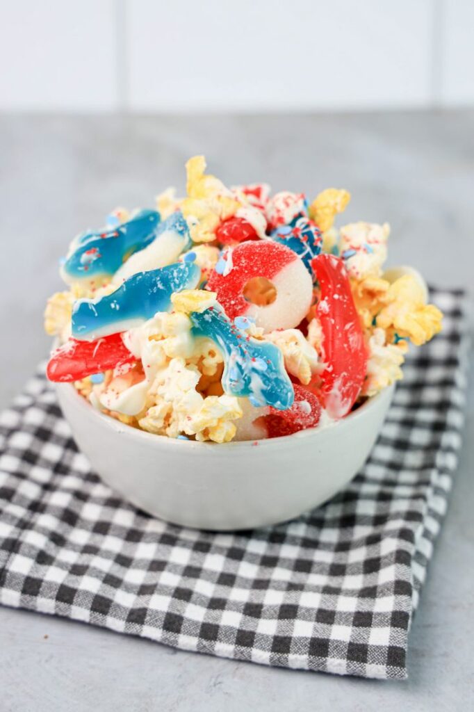 Shark week popcorn with shark gummies and red swedish fish in a white bowl on a gray plaid napkin on a concrete backdrop