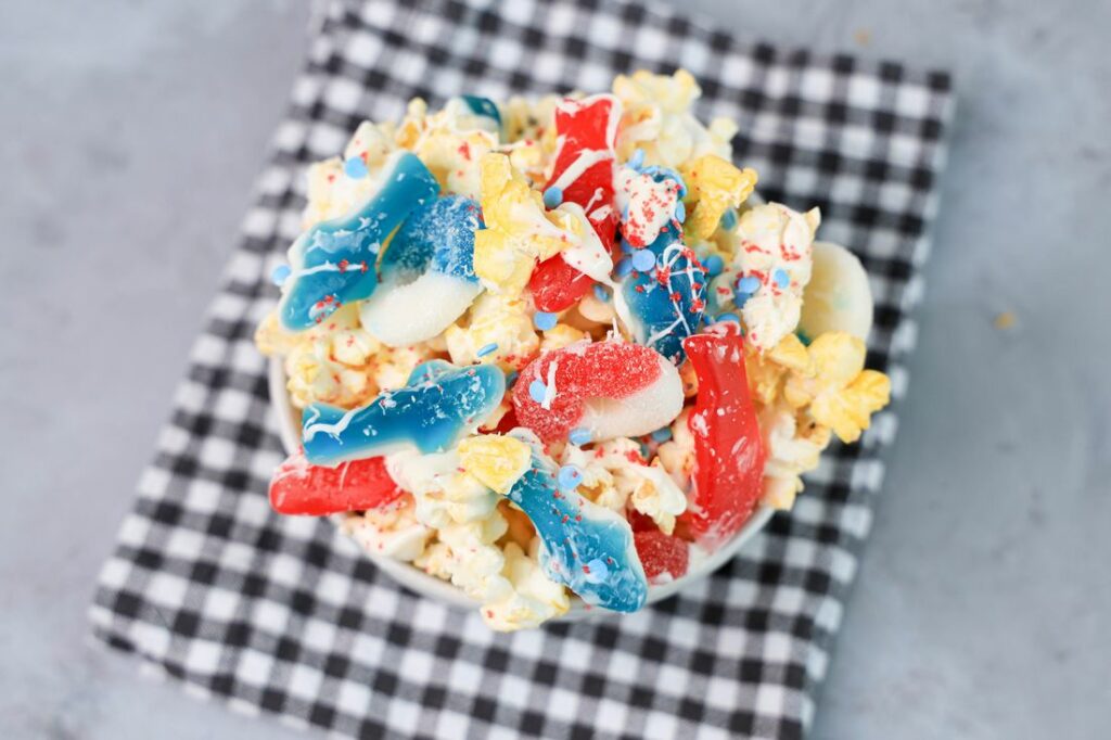 Shark week popcorn with shark gummies and red swedish fish in a white bowl on a gray plaid napkin on a concrete backdrop