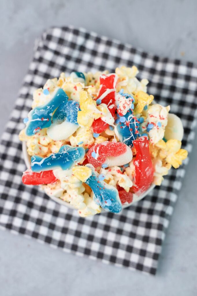 Shark week popcorn with shark gummies and red swedish fish in a white bowl on a gray plaid napkin on a concrete backdrop