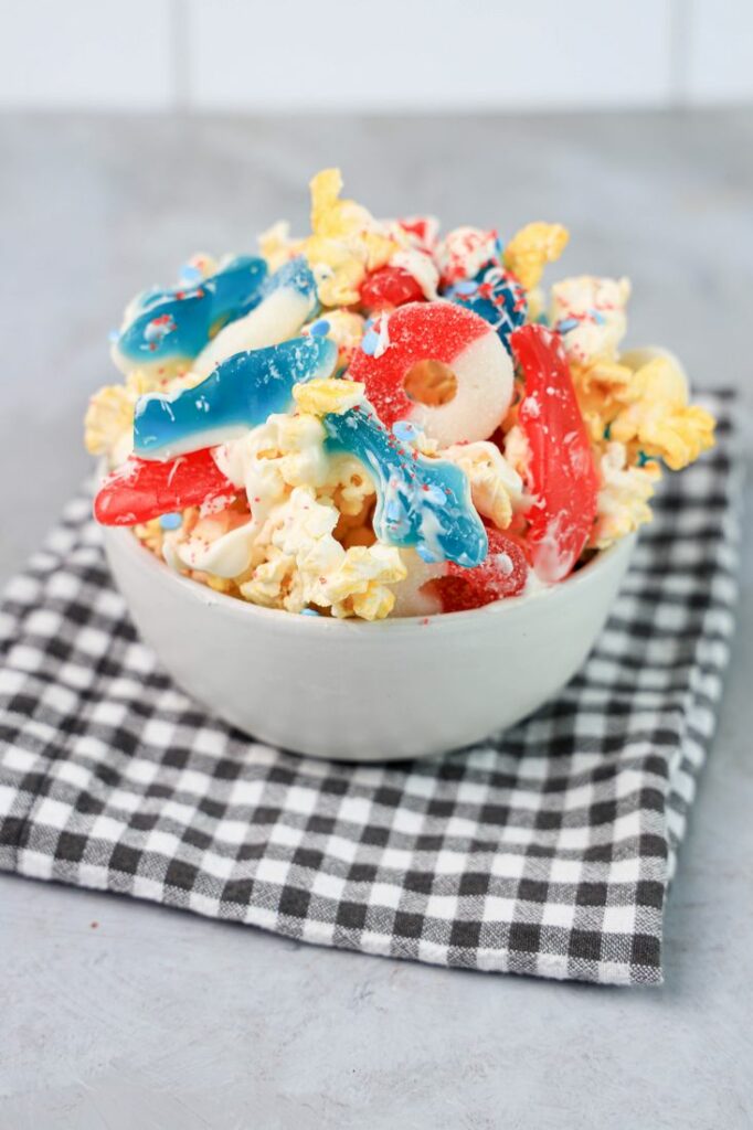 Shark week popcorn with shark gummies and red swedish fish in a white bowl on a gray plaid napkin on a concrete backdrop