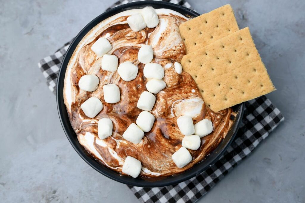 Smores dip with marshmallows in a black bowl on a gray plaid napkin with concrete backdrop