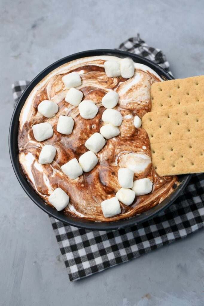 Smores dip with marshmallows in a black bowl on a gray plaid napkin with concrete backdrop