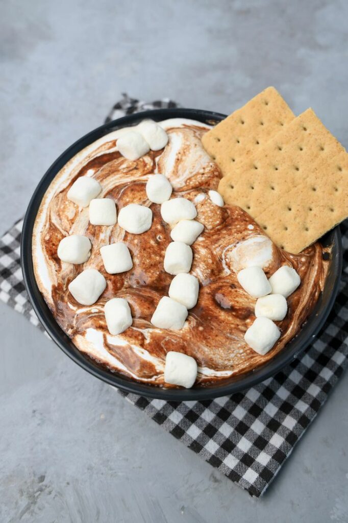 Smores dip with marshmallows in a black bowl on a gray plaid napkin with concrete backdrop