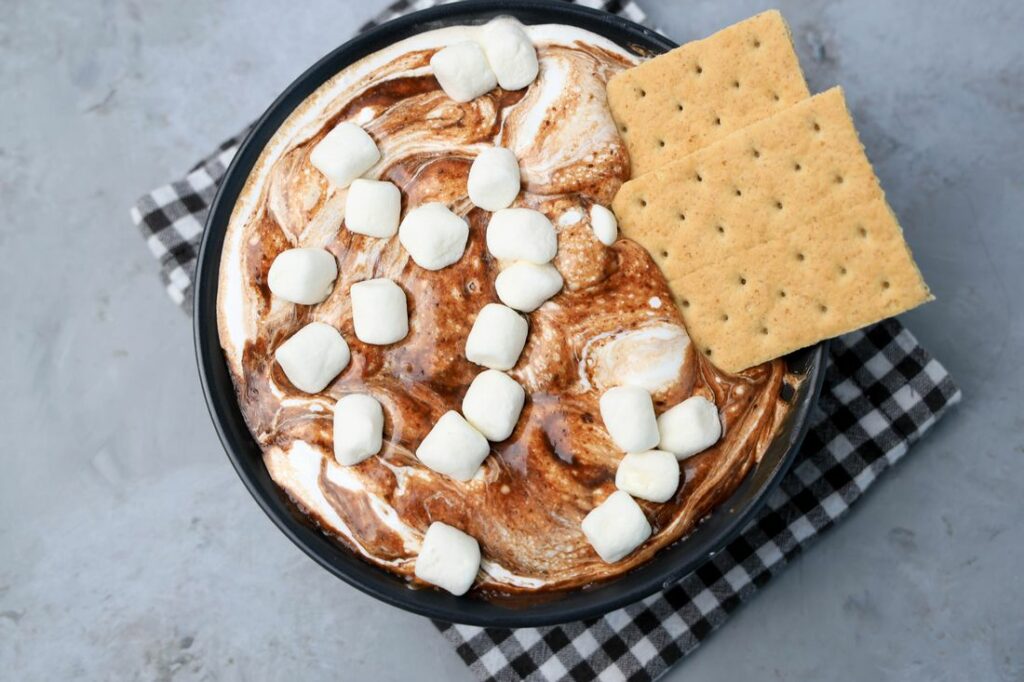 Smores dip with marshmallows in a black bowl on a gray plaid napkin with concrete backdrop