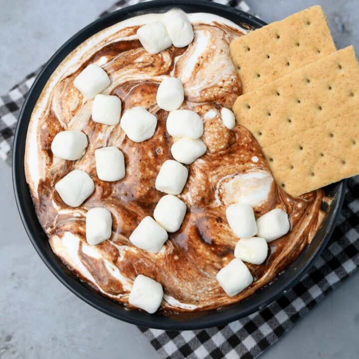 Smores dip with marshmallows in a black bowl on a gray plaid napkin with concrete backdrop