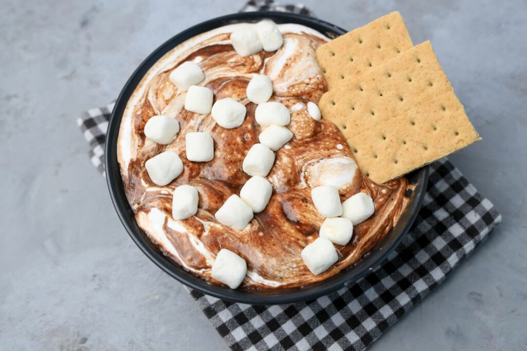 Smores dip with marshmallows in a black bowl on a gray plaid napkin with concrete backdrop