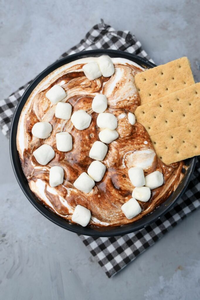 Smores dip with marshmallows in a black bowl on a gray plaid napkin with concrete backdrop