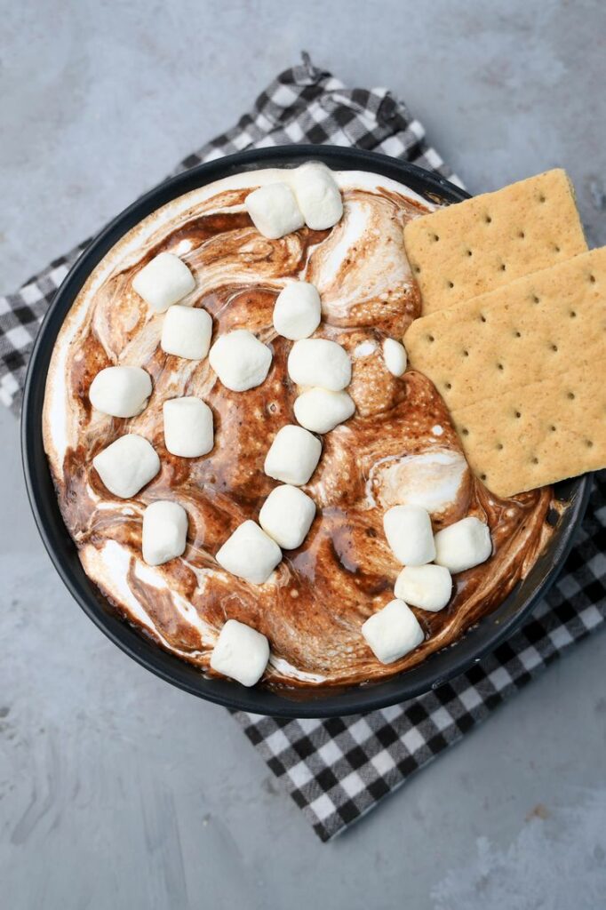 Smores dip with marshmallows in a black bowl on a gray plaid napkin with concrete backdrop