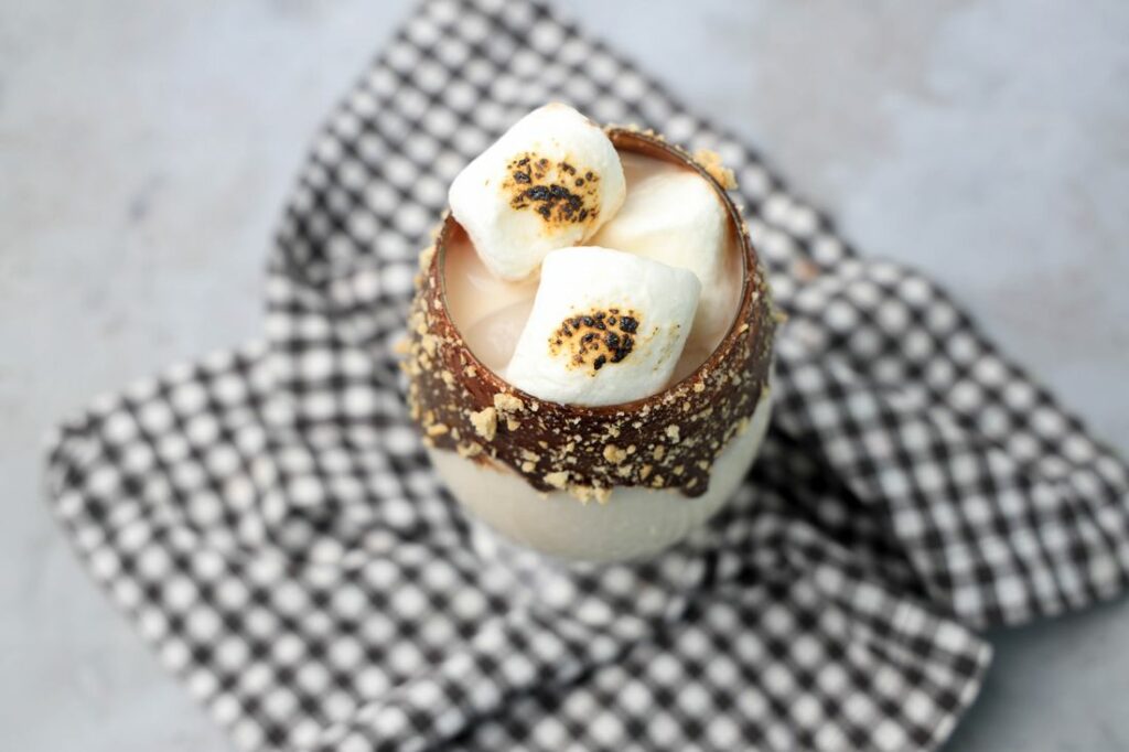 Campfire cocktail in a glass with chocolate and marshmallows on a plaid napkin with a concrete backdrop