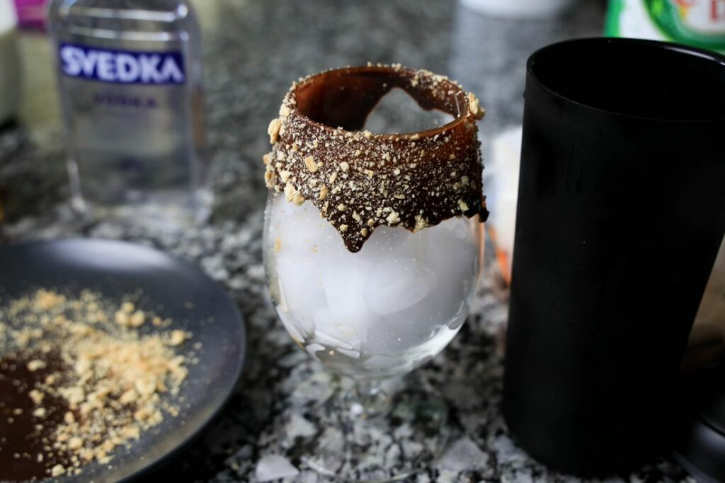 Glass with ice and chocolate syrup on the edge along with graham cracker crumbs.