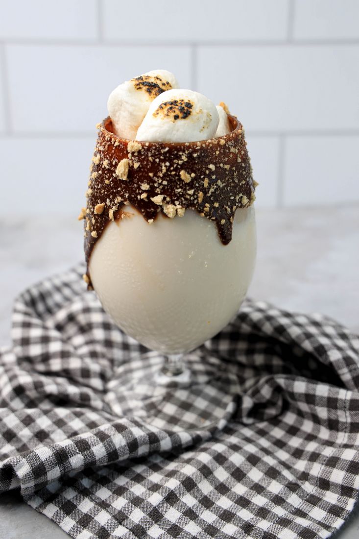 Campfire cocktail in a glass with chocolate and marshmallows on a plaid napkin with a concrete backdrop