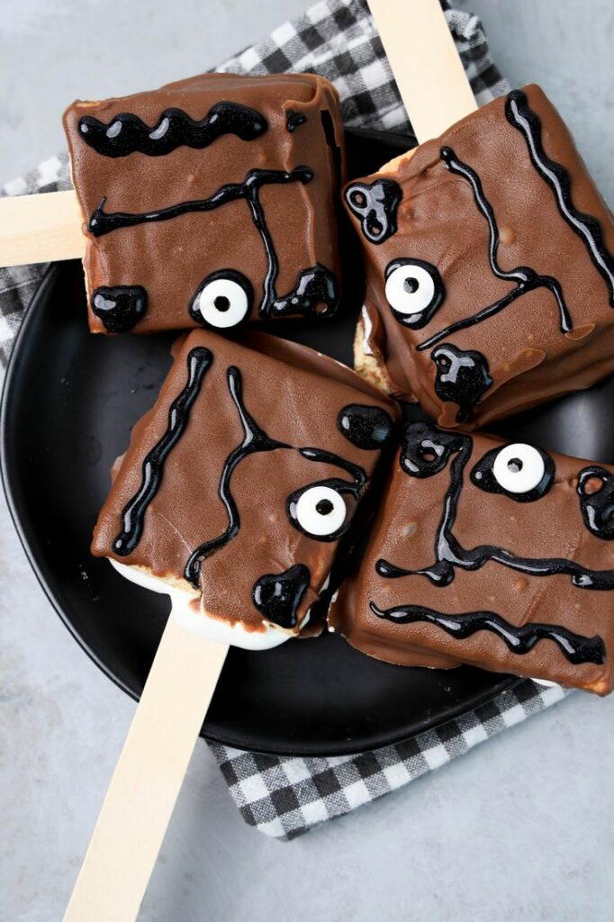 Hocus Pocus Book Of Spells S'mores Pops on a black plate with a gray plaid napkin on a faux concrete backdrop