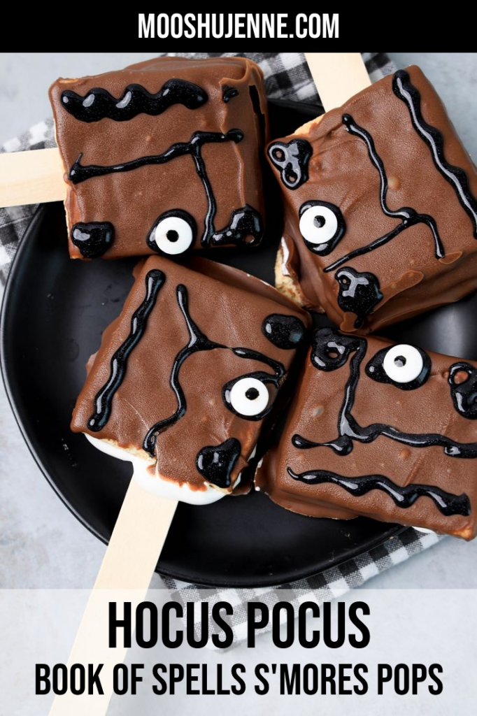 Hocus Pocus Book Of Spells S'mores Pops on a black plate with a gray plaid napkin on a faux concrete backdrop