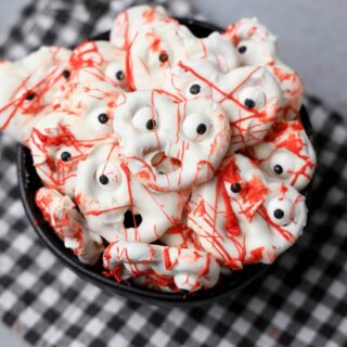 Bloody ghost pretzels in a black bowl with a gray plaid napkin on a faux concrete back drop.