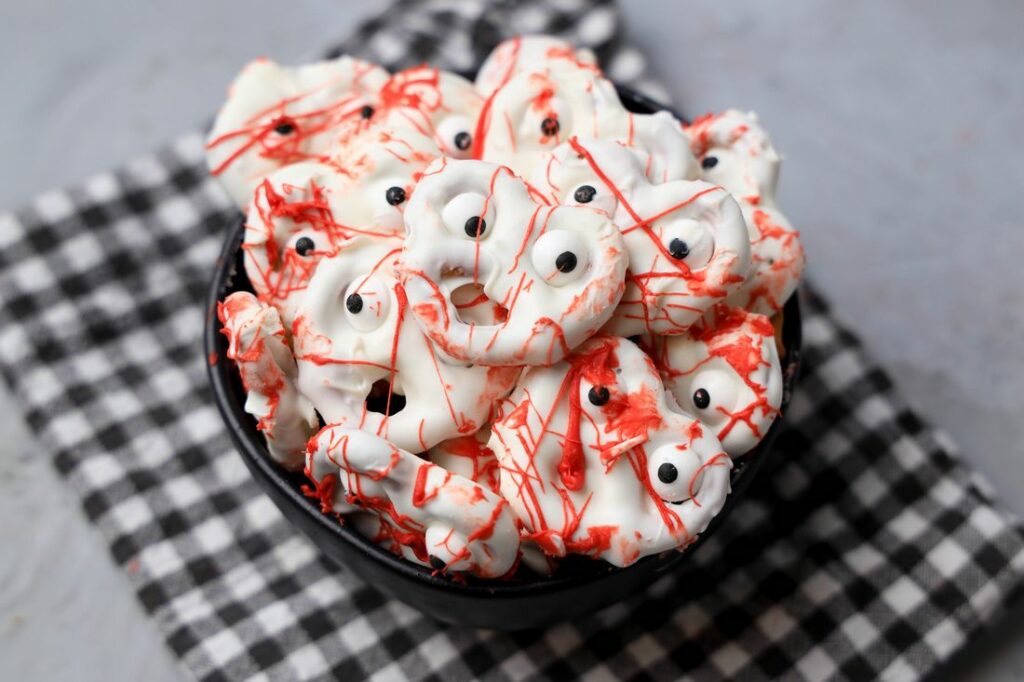 Bloody ghost pretzels in a black bowl with a gray plaid napkin on a faux concrete back drop.