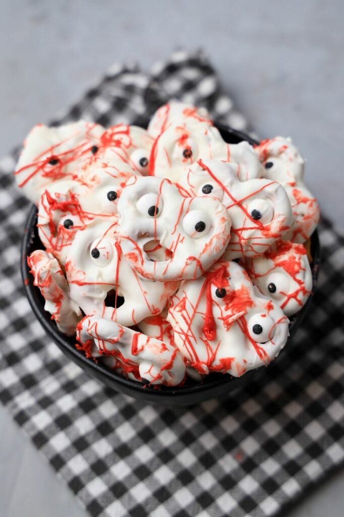 Bloody ghost pretzels in a black bowl with a gray plaid napkin on a faux concrete back drop.