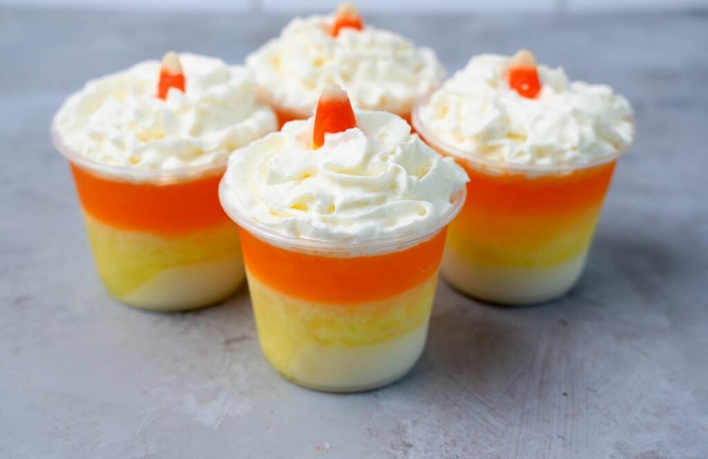 Candy corn layered jello shots topped with whipped topping and a piece of candy corn on a faux concrete backdrop