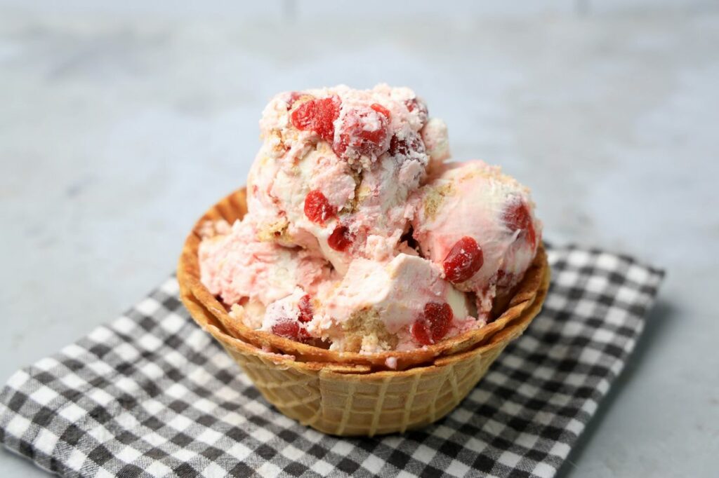 Cherry Pie Ice Cream in a waffle cone bowl with a gray plaid napkin on a faux concrete backdrop.