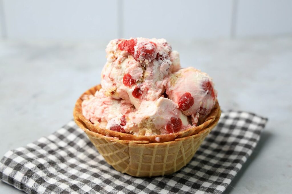 Cherry Pie Ice Cream in a waffle cone bowl with a gray plaid napkin on a faux concrete backdrop.