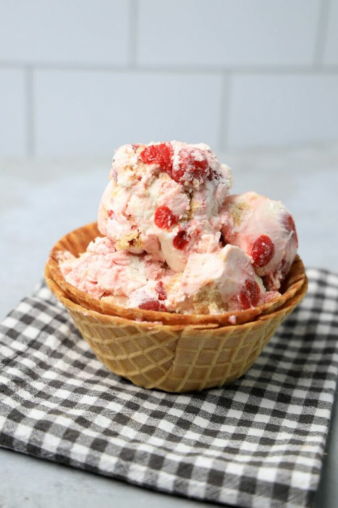 Cherry Pie Ice Cream in a waffle cone bowl with a gray plaid napkin on a faux concrete backdrop.