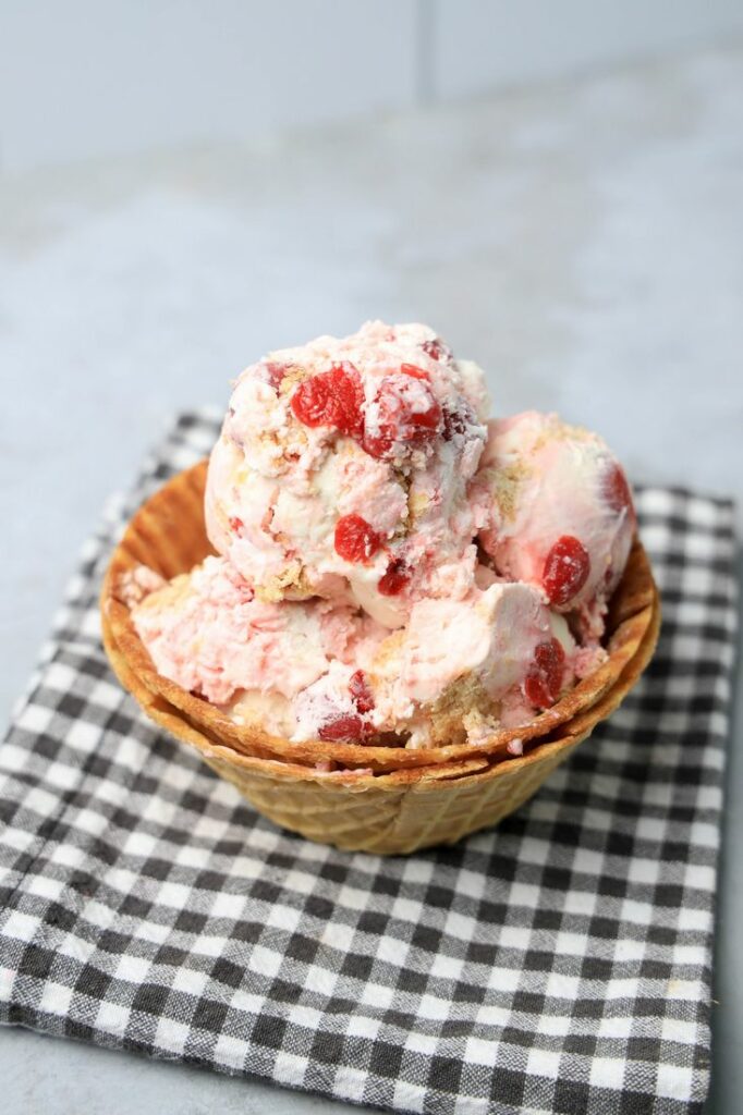 Cherry Pie Ice Cream in a waffle cone bowl with a gray plaid napkin on a faux concrete backdrop.