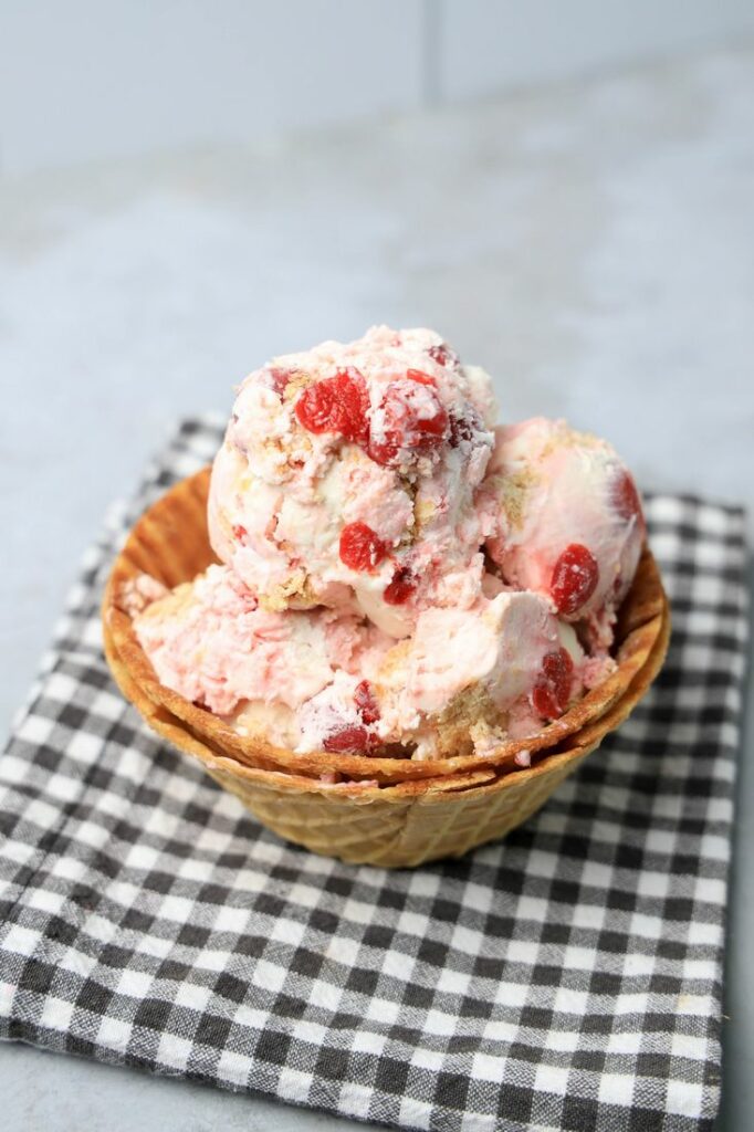 Cherry Pie Ice Cream in a waffle cone bowl with a gray plaid napkin on a faux concrete backdrop.