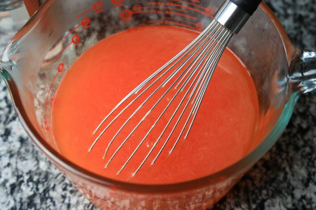 Orange jello in a glass bowl with a whisk.