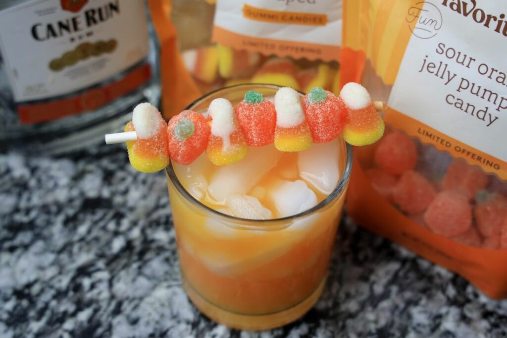 Glass with ice along with fantas soda, orange juice, gummy candy corns, and gummy pumpkins.