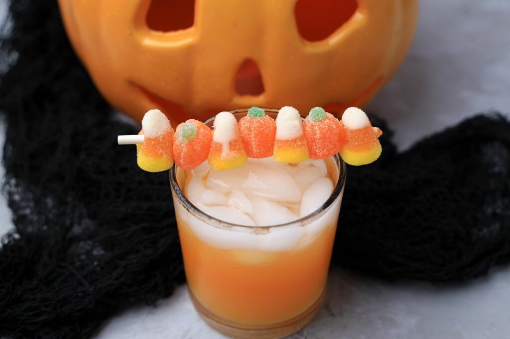 Halloween Michael Myers Cocktail with orange juice and orange soda. Topped with candy corn and pumpkin gummies. With the original myer's pumpkin in the back.