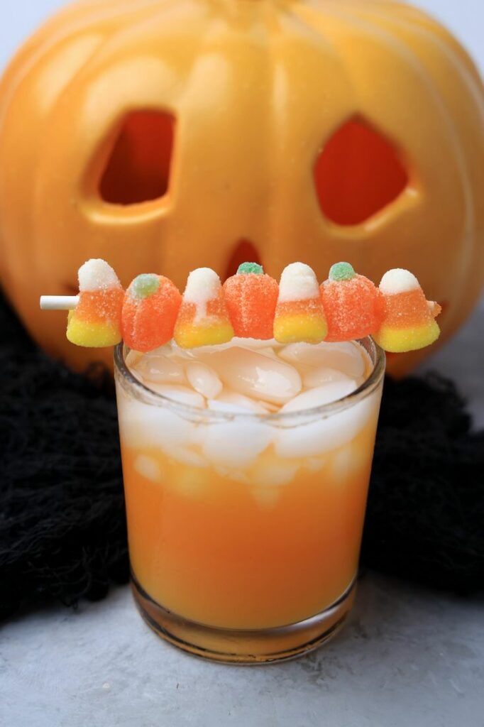 Halloween Michael Myers Cocktail with orange juice and orange soda. Topped with candy corn and pumpkin gummies. With the original myer's pumpkin in the back.