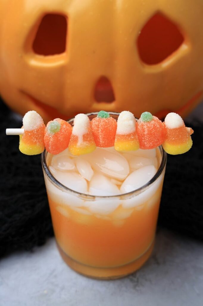 Halloween Michael Myers Cocktail with orange juice and orange soda. Topped with candy corn and pumpkin gummies. With the original myer's pumpkin in the back.