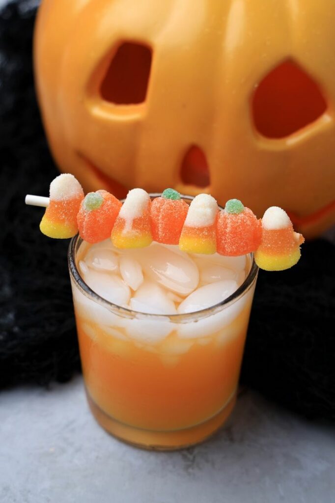 Halloween Michael Myers Cocktail with orange juice and orange soda. Topped with candy corn and pumpkin gummies. With the original myer's pumpkin in the back.