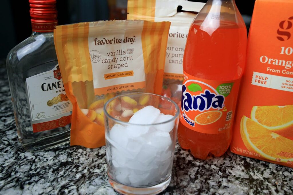 Glass with ice along with fantas soda, orange juice, gummy candy corns, and gummy pumpkins.