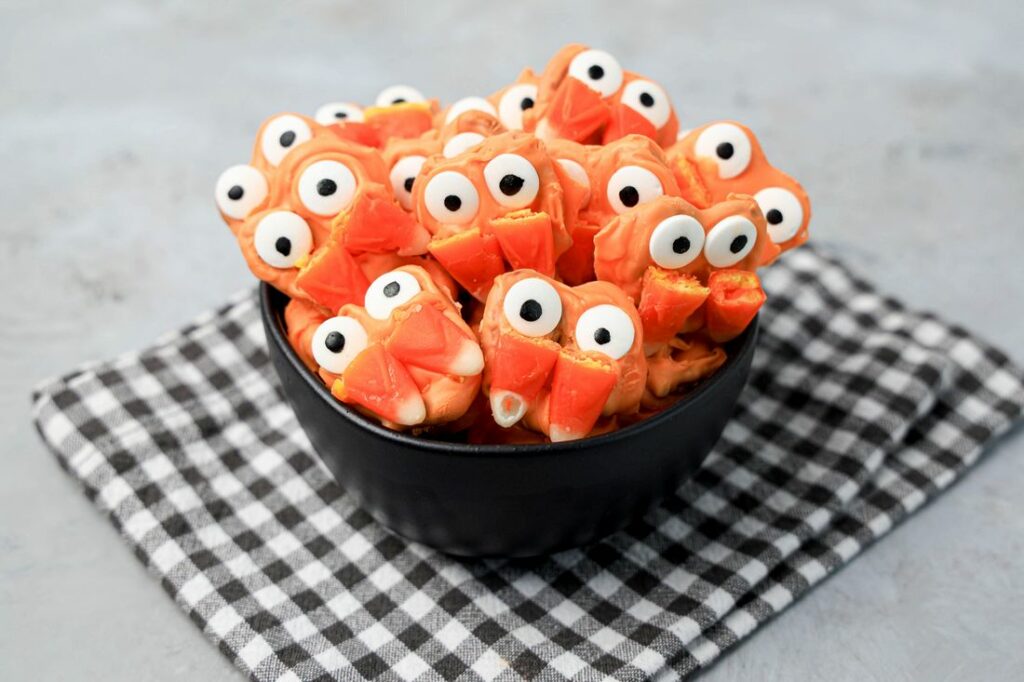 Pretzels with candy eyes and candy corn as fangs. In a black bowl on a plaid napkin.