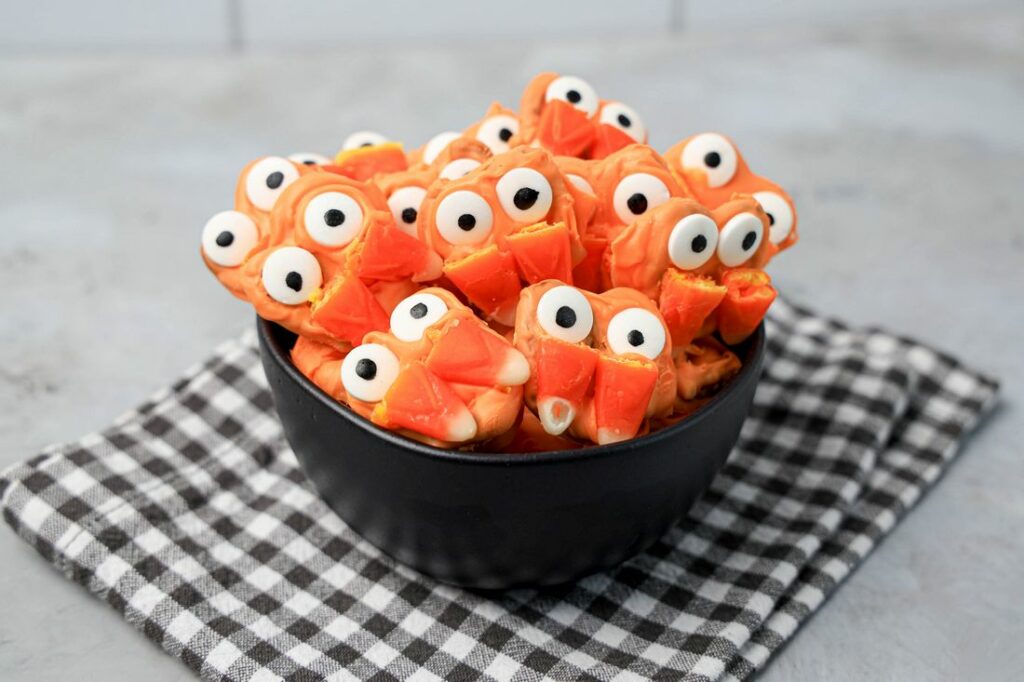 Pretzels with candy eyes and candy corn as fangs. In a black bowl on a plaid napkin.