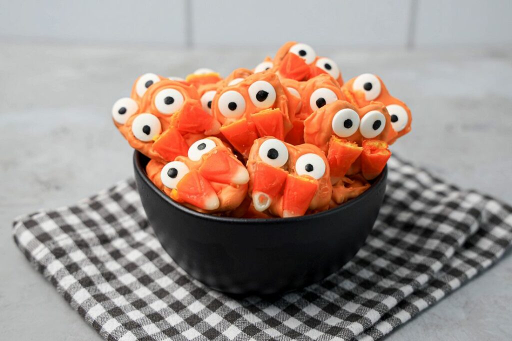 Pretzels with candy eyes and candy corn as fangs. In a black bowl on a plaid napkin.