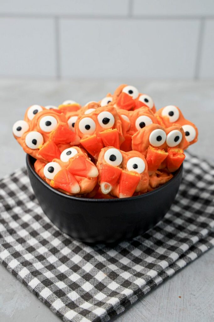 Pretzels with candy eyes and candy corn as fangs. In a black bowl on a plaid napkin.