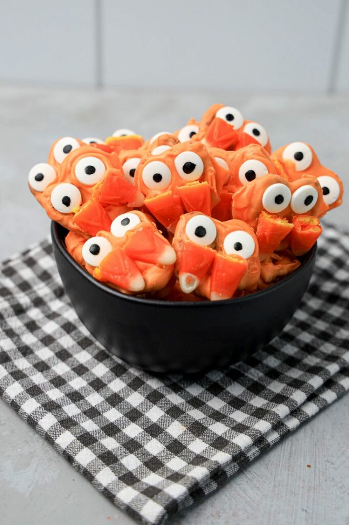 Pretzels with candy eyes and candy corn as fangs. In a black bowl on a plaid napkin.