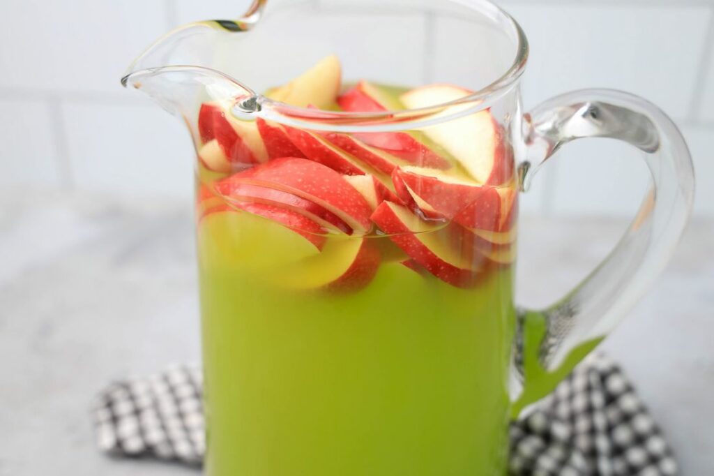 Grinch Christmas Punch with red apples in a glass pitcher on a gray plaid napkin.